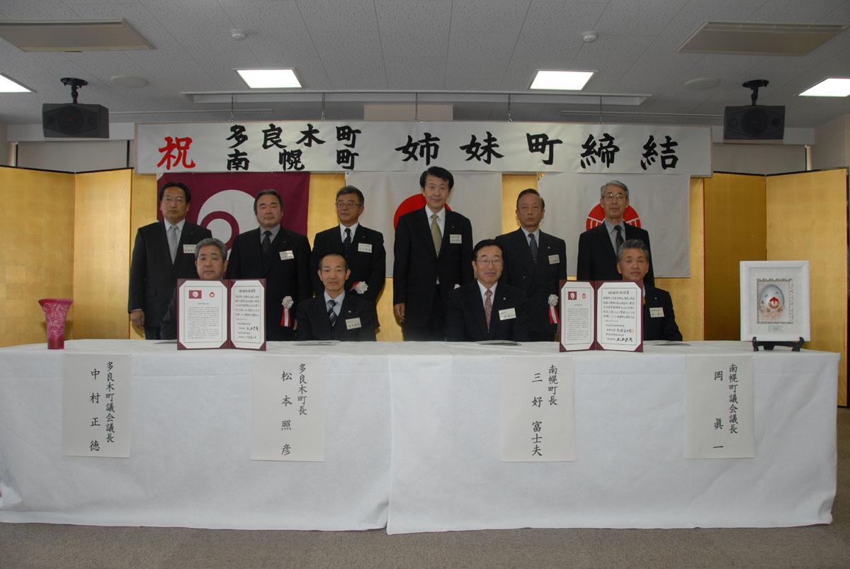 多良木町と北海道南幌町の姉妹町締結の様子を写した写真。後ろに飾られた金屏風の前方にある長机には双方の町の町長、議会議長が1人ずつ座っており、その後ろに6名の関係者が並んで立っています。